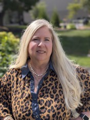 Headshot of Lisa Mansfield smiling outdoors with lush green trees and foliage in the soft-focus background. She has long, blonde hair and is wearing a leopard-print button-up shirt with a denim collar and layered necklaces.