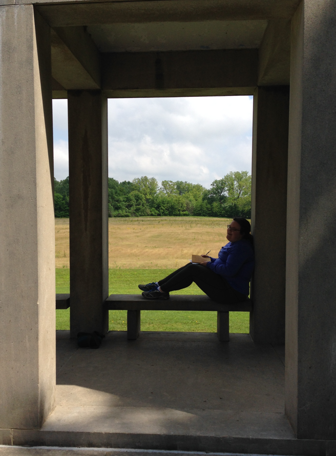 A student sits and writes with nature at her backdrop as she sits in a square concrete coliseum.